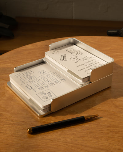 The Complete Analog Collection (Silver Aluminum) neatly holds stacked note cards with sketches and notes on a wooden desk beside a black and gold pen, while warm light casts gentle shadows across the scene.
