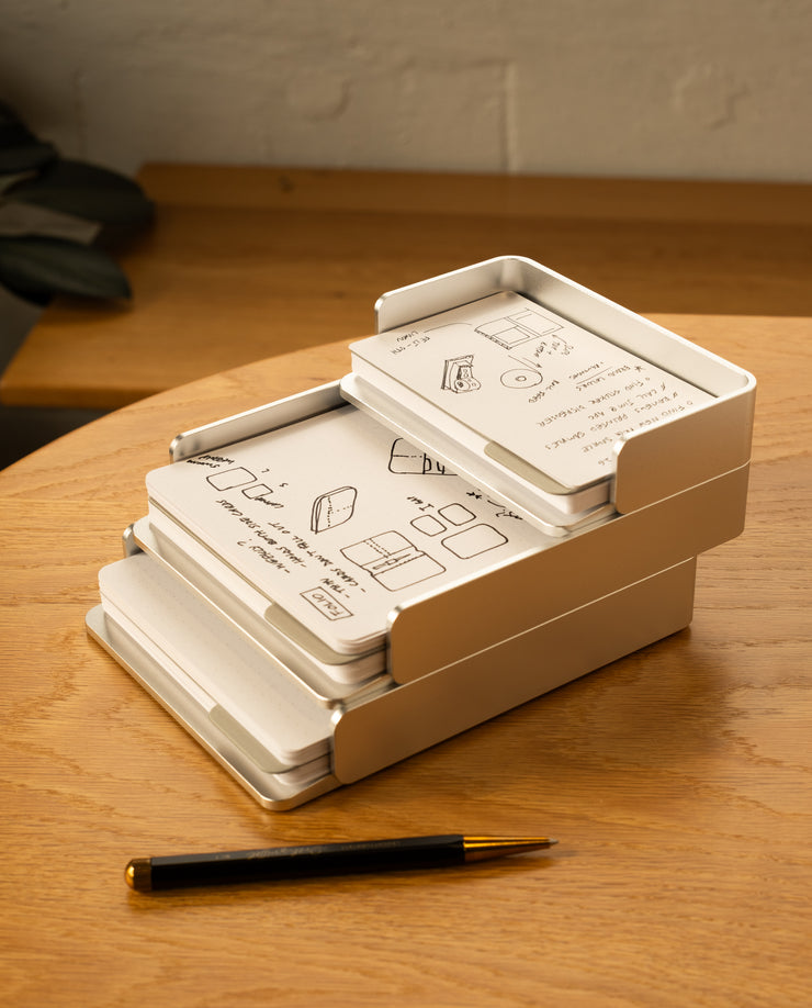 The Analog Capture Kit - 2 Large (Silver Aluminum) sits on a wooden desk, holding illustrated instruction cards. A black and gold pen is nearby, with part of a gray item and a light-colored wall in the background.