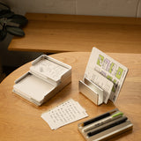 A wooden table displays The Complete Analog Collection (Silver Aluminum), organizing notecards, pens, and markers. Some cards show handwritten notes and sketches, with a plant partially visible in the background.