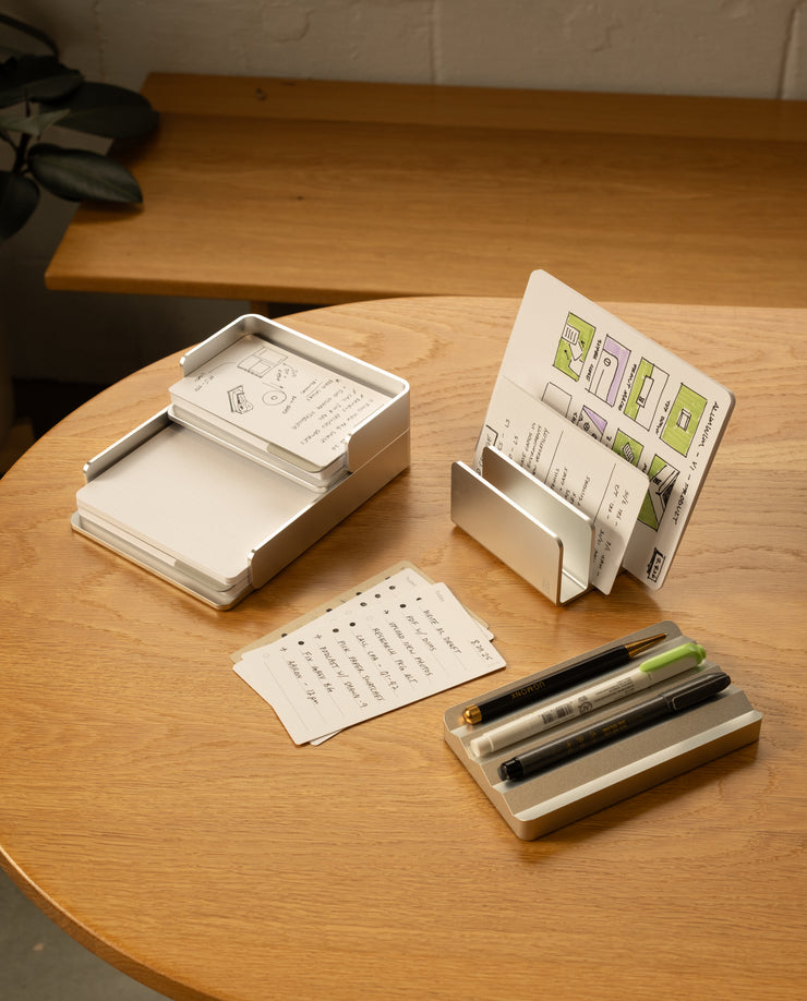 A wooden table displays The Complete Analog Collection (Silver Aluminum), organizing notecards, pens, and markers. Some cards show handwritten notes and sketches, with a plant partially visible in the background.