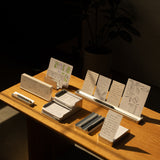The Complete Analog Collection (Silver Aluminum) is displayed on a wooden table with neatly arranged pens, notepads, and a card organizer holding sketched white cards, all bathed in warm sunlight with a potted plant and dark furniture in the background.