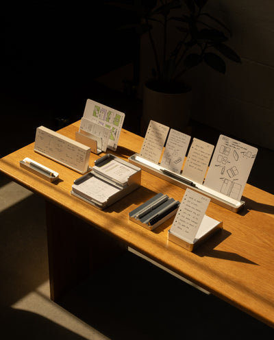 The Complete Analog Collection (Silver Aluminum) is displayed on a wooden table with neatly arranged pens, notepads, and a card organizer holding sketched white cards, all bathed in warm sunlight with a potted plant and dark furniture in the background.
