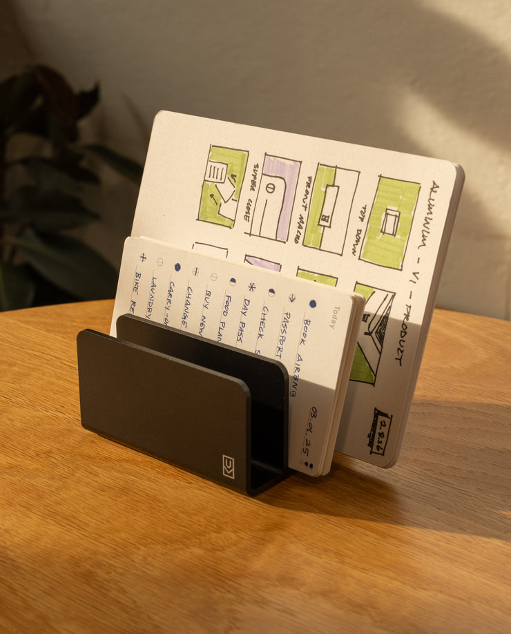 The Complete Analog Collection (Black Aluminum) holds two open notebooks on a wooden desk, one featuring colorful diagrams and the other with handwritten notes. Sunlight from a window casts warm shadows across the organized workspace.