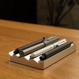 A close-up of three pens from The Complete Analog Collection (Silver Aluminum) resting in a white holder on a wooden desk, bringing elegant style to your desktop organizer.