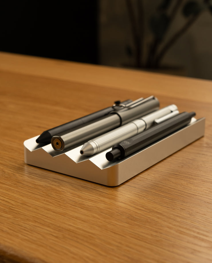 A close-up of three pens from The Complete Analog Collection (Silver Aluminum) resting in a white holder on a wooden desk, bringing elegant style to your desktop organizer.