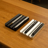 Two Analog Multi Pen Trays in black and silver hold three pens each, side by side on a wooden surface. These solid aluminum trays feature grooves to keep pens organized.