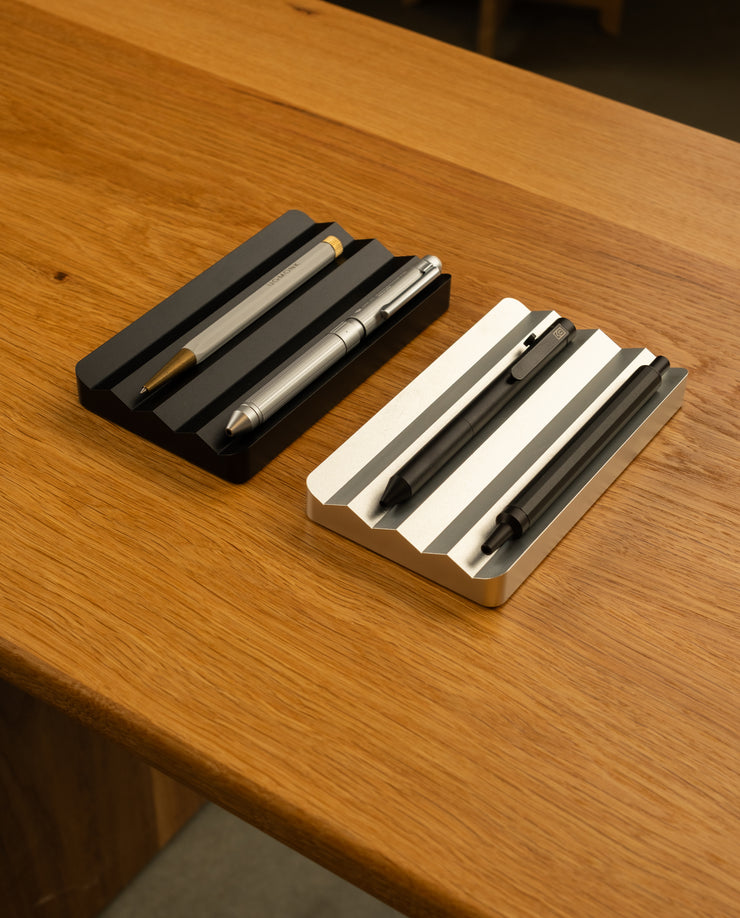 Two Analog Multi Pen Trays in black and silver hold three pens each, side by side on a wooden surface. These solid aluminum trays feature grooves to keep pens organized.
