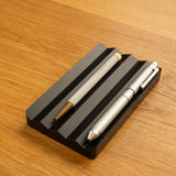 The Complete Analog Collection (Black Aluminum) features two metallic pens resting parallel on a black grooved holder atop a light wooden surface—a sleek, modern desktop organizer for your workspace.