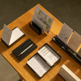 A wooden table displays The Complete Analog Collection (Black Aluminum), organizing notepads, pens, and sketches. The well-lit scene features a plain gray floor in the background.