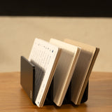 The Analog Card Organizer in black aluminum displays several open notebooks with lined and blank pages on a wooden surface against a light-colored wall.
