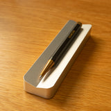 A black and gold pen rests diagonally on a rectangular gray and white holder from The Complete Analog Collection (Silver Aluminum), set atop a light wooden surface.