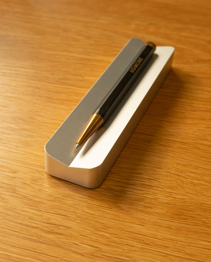 A black and gold pen rests diagonally on a rectangular gray and white holder from The Complete Analog Collection (Silver Aluminum), set atop a light wooden surface.