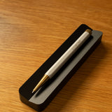 A pen from The Complete Analog Collection (Black Aluminum) rests diagonally on a black rectangular holder atop a warm wooden surface—perfect for enhancing any desktop organizer setup.