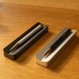 A silver aluminum Analog Pen Tray holds two stylish pens—one silver, one black with gold accents—creating a sleek desk accessory atop a wooden surface.