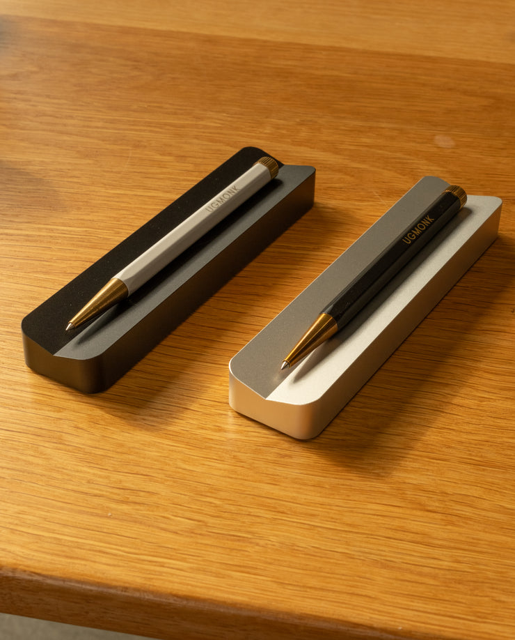 A silver aluminum Analog Pen Tray holds two stylish pens—one silver, one black with gold accents—creating a sleek desk accessory atop a wooden surface.