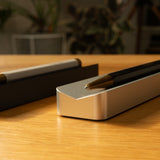 A close-up of two pens on Analog Pen Tray (Silver Aluminum)—a modern desk accessory on a wooden surface, with blurred indoor plants in the background.