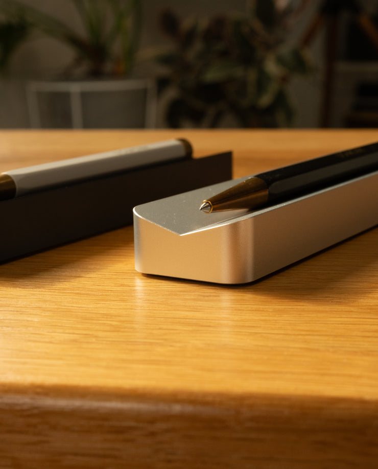 A close-up of two pens on Analog Pen Tray (Silver Aluminum)—a modern desk accessory on a wooden surface, with blurred indoor plants in the background.