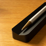 A close-up of a white and gold pen resting on the Analog Pen Tray (Black Aluminum), placed on a wooden surface.