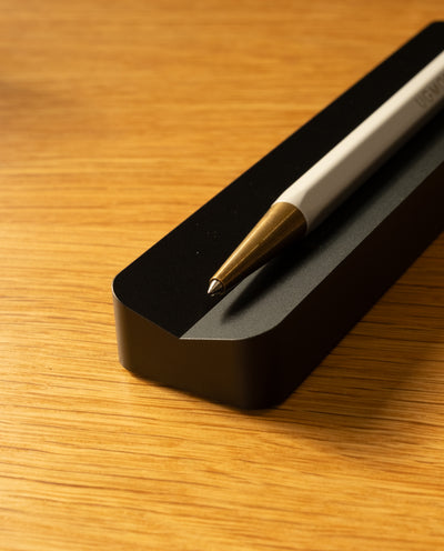 A close-up of a white and gold pen resting on the Analog Pen Tray (Black Aluminum), placed on a wooden surface.