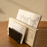 The Analog Card Organizer (Black Aluminum) holds multiple open notebooks with handwritten notes and lists, all neatly arranged upright on a wooden table bathed in warm light.