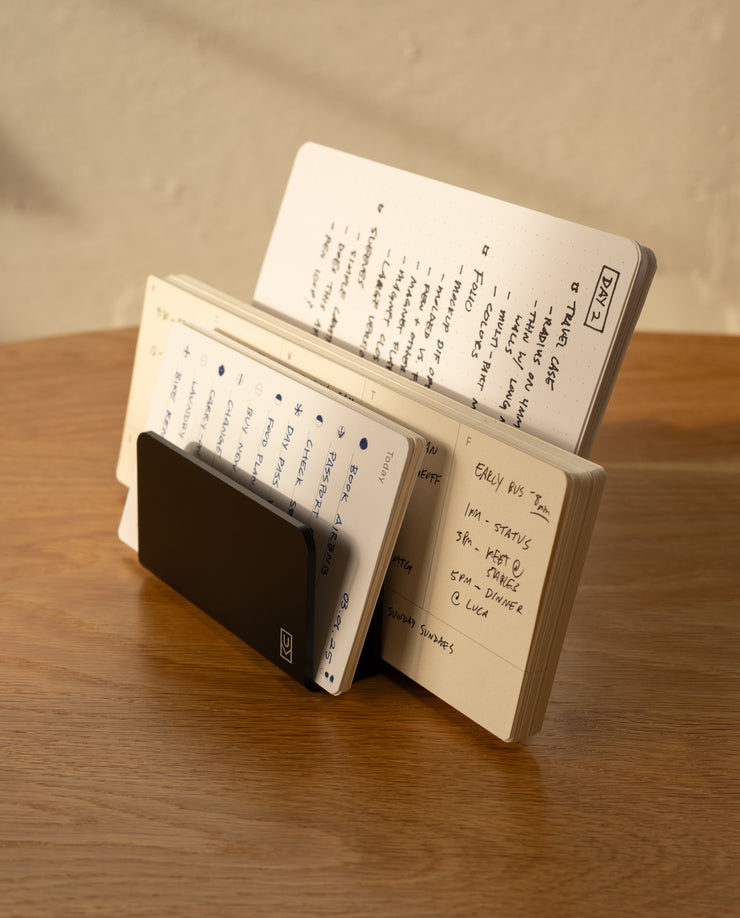 The Analog Card Organizer (Black Aluminum) holds multiple open notebooks with handwritten notes and lists, all neatly arranged upright on a wooden table bathed in warm light.