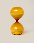 The 15 Minute Hourglass (Amber) features a transparent orange design filled with yellow sand and sits elegantly on a plain light background.