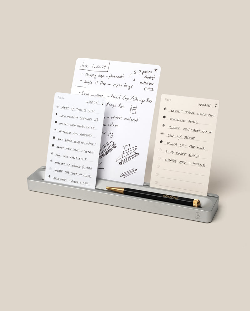 A metal desktop organizer holds three note cards with handwritten lists and sketches, along with a black and gold pen resting in a groove at the front. The background is a plain light beige.
