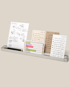 A minimalist metal desk organizer holds four notecards with hand-drawn sketches, lists, and a color-coded chart, along with a black pen and pencil, all displayed on a neutral background.