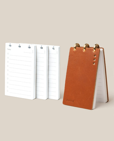 The Leather Discbound Analog + 3 Refills (Tan) features a tan full-grain leather notepad with brass rings and three stacks of white daily planner refills, all displayed on a beige background.