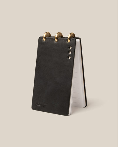 The Leather Discbound Analog (Black) is a compact notepad with full-grain leather covers, three large brass binding rings, and silver rivets on the front. It stands upright, opened to lined pages—ideal for Analog Task Cards.