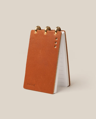 The Leather Discbound Analog (Tan) stands upright on a beige background, featuring full-grain tan leather covers, brass rings, four metal rivets, and lined pages ideal for Discbound Analog Task Cards.