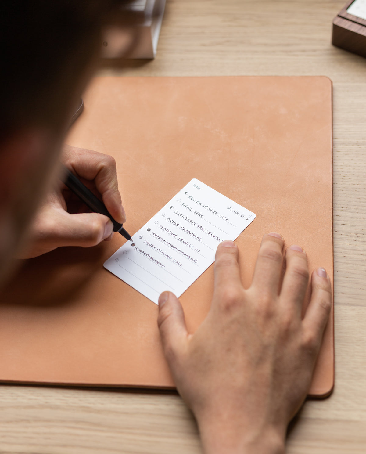 A person writes on an Analog - Today Card placed on a tan desk mat, using the card for effective task prioritization. The Analog - Today Card includes tasks such as cleaning, breakfast, and phone calls.
