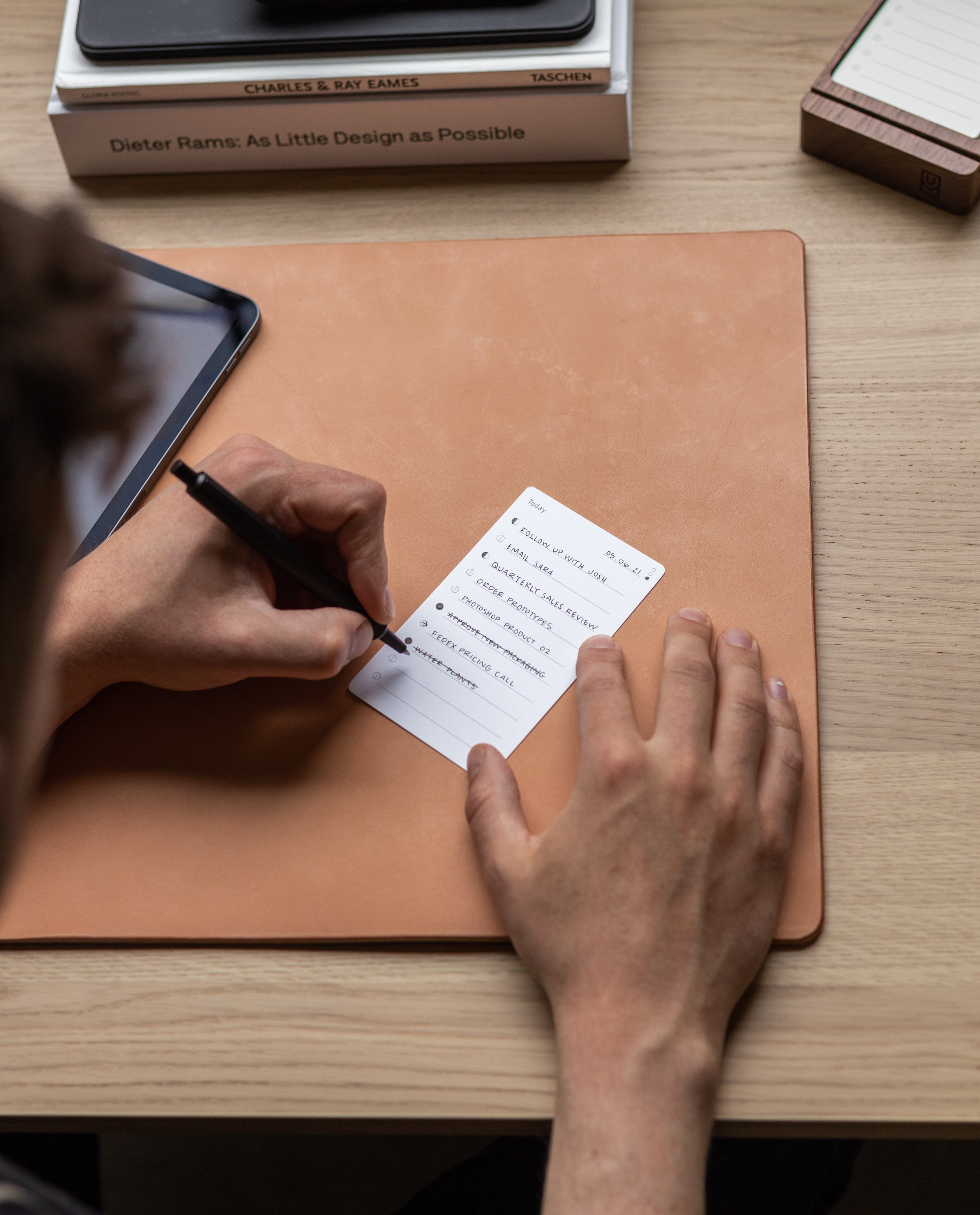 Someone writes a to-do list on an Analog Annual Refill card while sitting at a desk with a leather mat, tablet, and stacked books, including Dieter Rams: As Little Design as Possible.