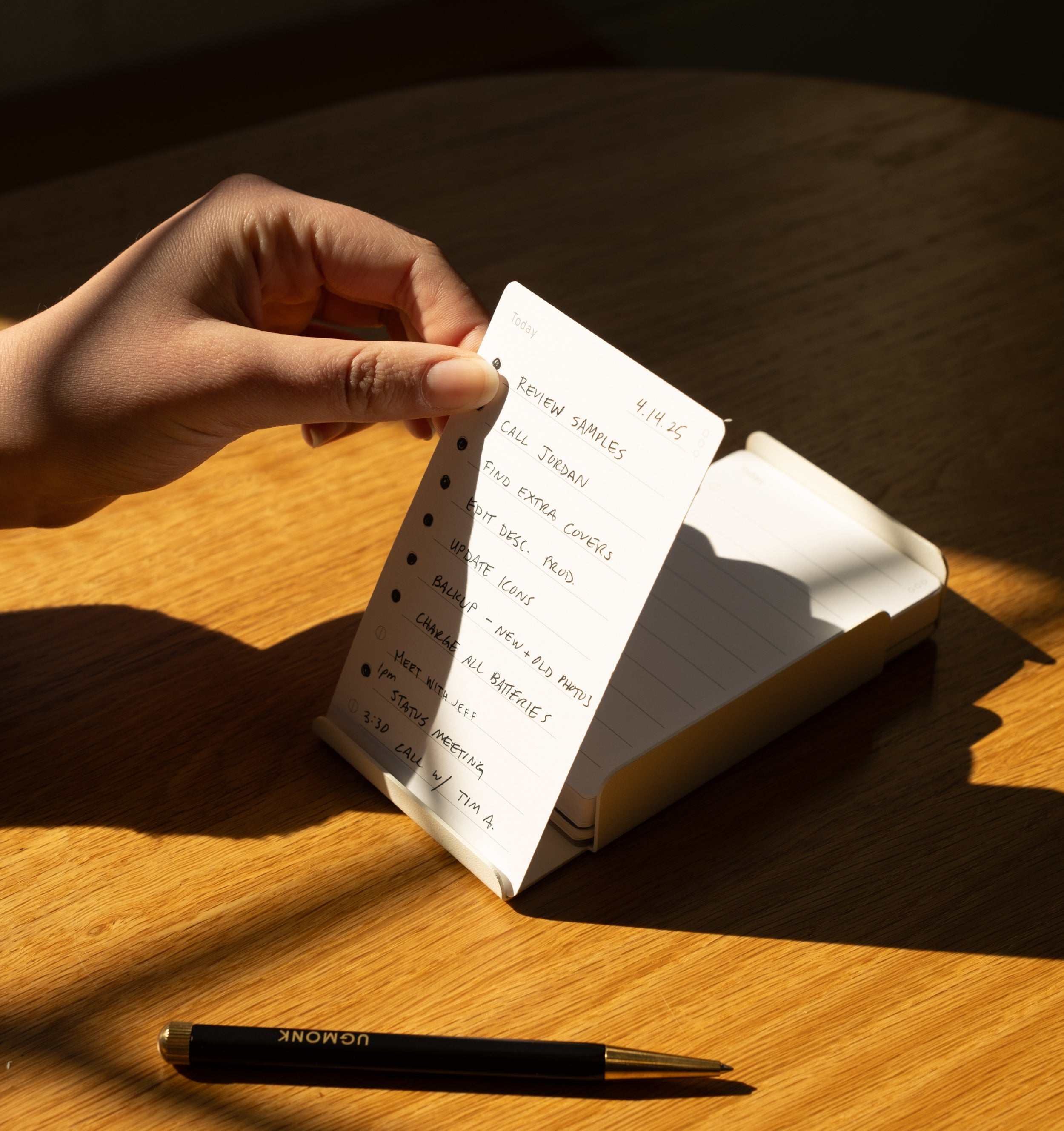 A hand holds a small notepad with a handwritten to-do list on a wooden table; a black pen labeled UGMONK rests nearby, and sunlight casts shadows across the scene.