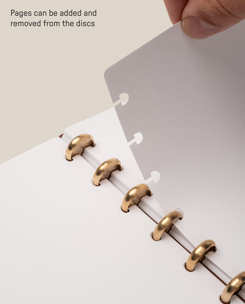 A hand removes a sheet of blank journal paper from a Discbound Journal with gold discs using the Discbound Journal Refill - Blank (Standard 6″ x 8.5″). Text reads, Pages can be added and removed from the discs, making it easy to organize or use refill paper.