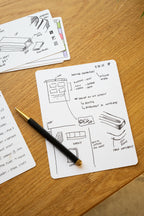 A notecard on a wooden desk displays hand-drawn sketches, notes, and a to-do list, with a black and gold pen resting on top. Another notecard with additional notes and sketches is partially visible in the background.