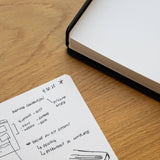 A black and gold pen rests on Analog Dot Grid Cards - Large with handwritten notes and sketches, while an open, blank dotted notebook lies nearby on a wooden desk.