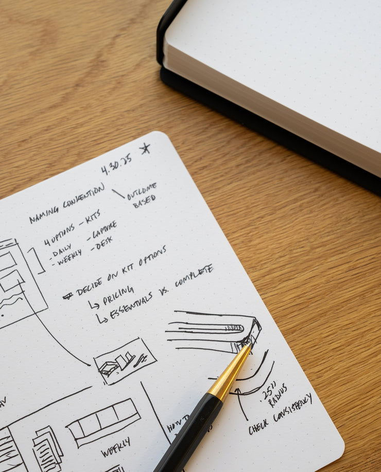 A black and gold pen rests on Analog Dot Grid Cards - Large with handwritten notes and sketches, while an open, blank dotted notebook lies nearby on a wooden desk.