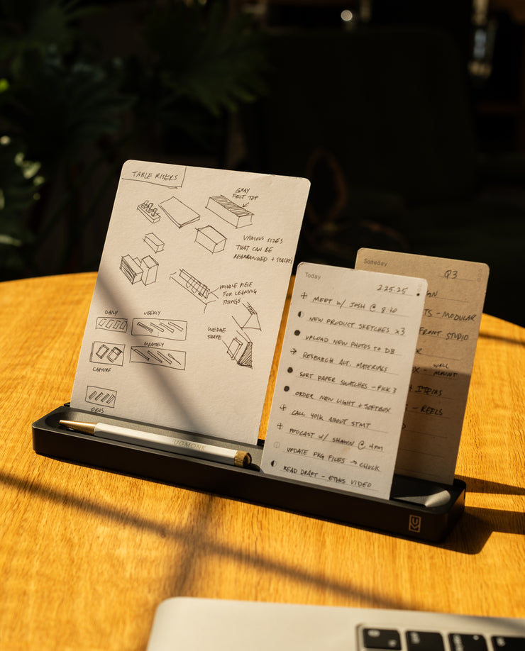 A notepad with hand-drawn sketches and a to-do list rests on a wooden table, held upright by a black stand with a pen. Sunlight shines on the desk, and part of a laptop keyboard is visible in the foreground.