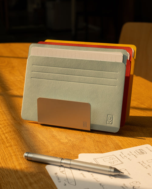 A metal pen holder displays colored index cards in Analog Card Sleeves - Large (Bright) on a wooden table, with a silver pen and a sheet of paper featuring sketches and handwritten notes nearby, all in warm sunlight.