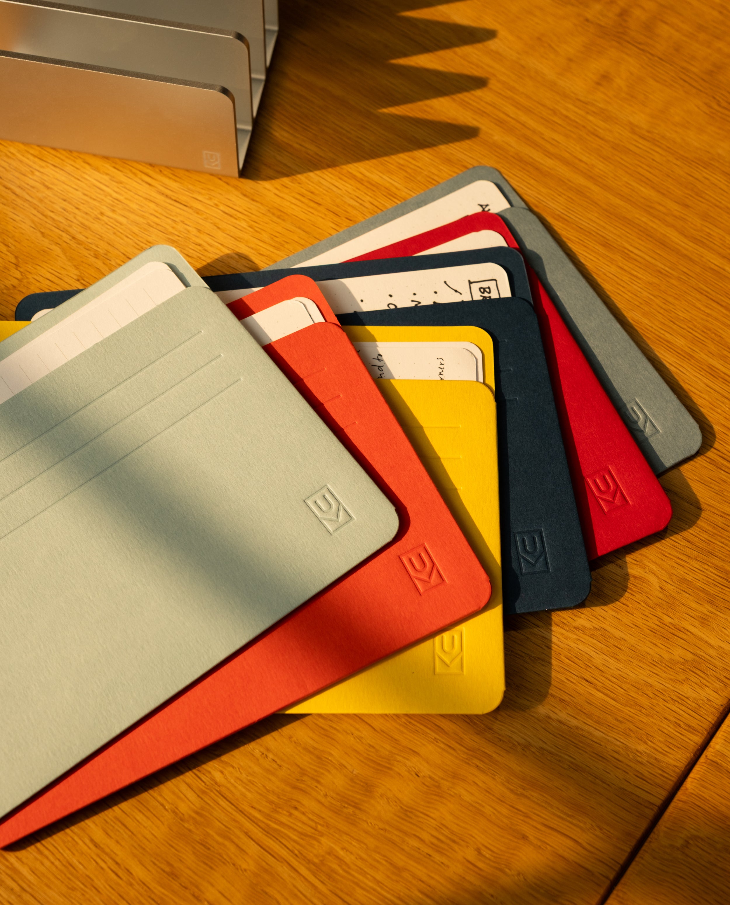 Assorted colorful notebooks and folders, partially stacked on a wooden surface, with some cards and papers tucked inside Analog Card Sleeves - Large (Bright). Sunlight casts shadows across the scene, highlighting the neat arrangement that doubles as a practical card organizer.