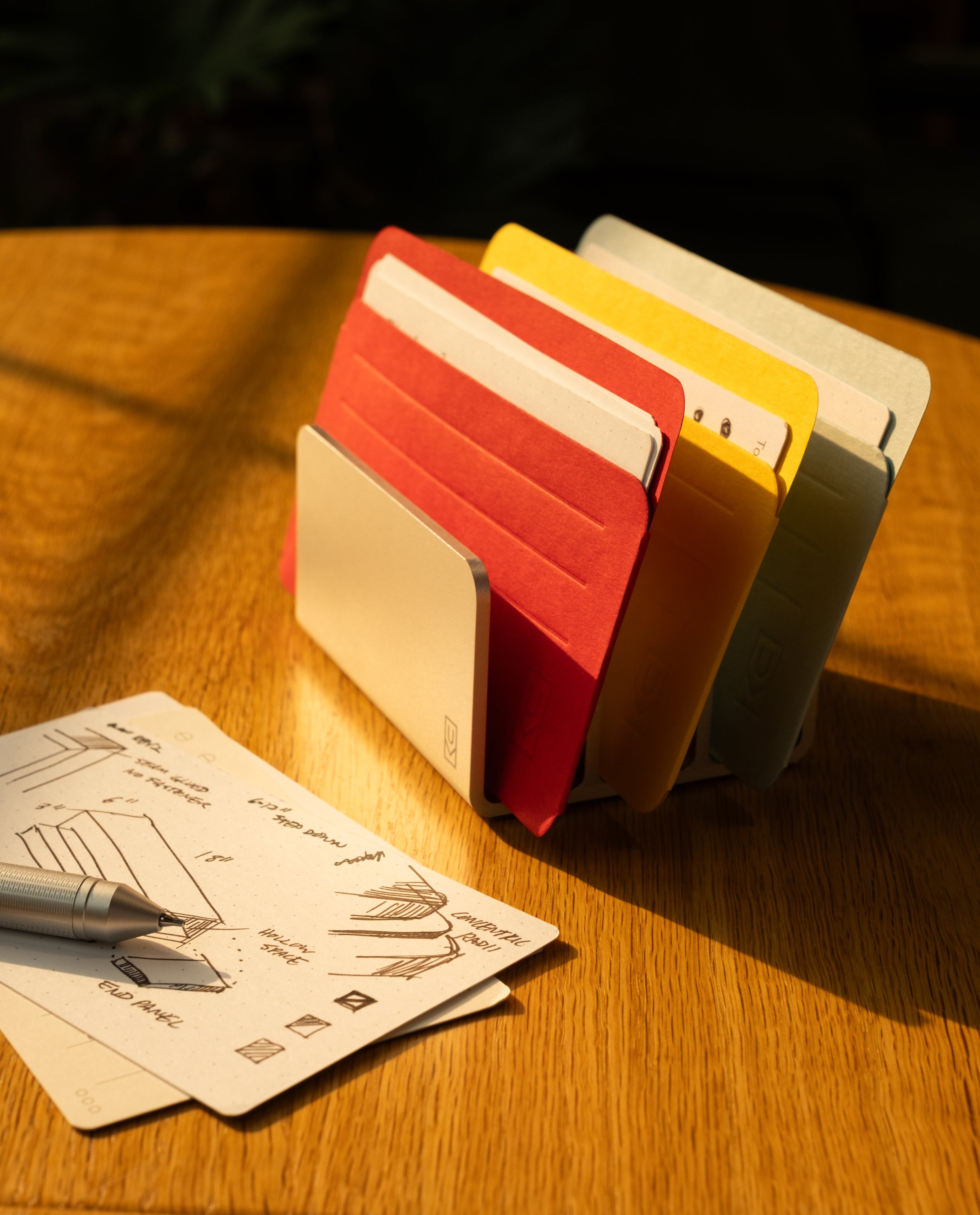 A metal card holder displaying colorful index cards in Analog Card Sleeves - Small (Bright) sits on a wooden table, while a pen rests on a few sketch-filled notecards nearby in warm sunlight.