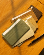 Five slim, minimalist Analog Card Sleeves - Small (Earth) in assorted earth tones are arranged on a wooden surface, each securely holding several cards. A black and gold pen rests nearby as sunlight casts gentle shadows over the scene, accentuating the refined design of these card organizers.