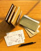 Three Analog Card Sleeves - Small (Earth) in brown, tan, and gray rest on a wooden surface beside loose notecards. One notecard displays hand-drawn woodworking diagrams, while a black and gold pen lies in front. Warm sunlight casts gentle shadows across the scene.