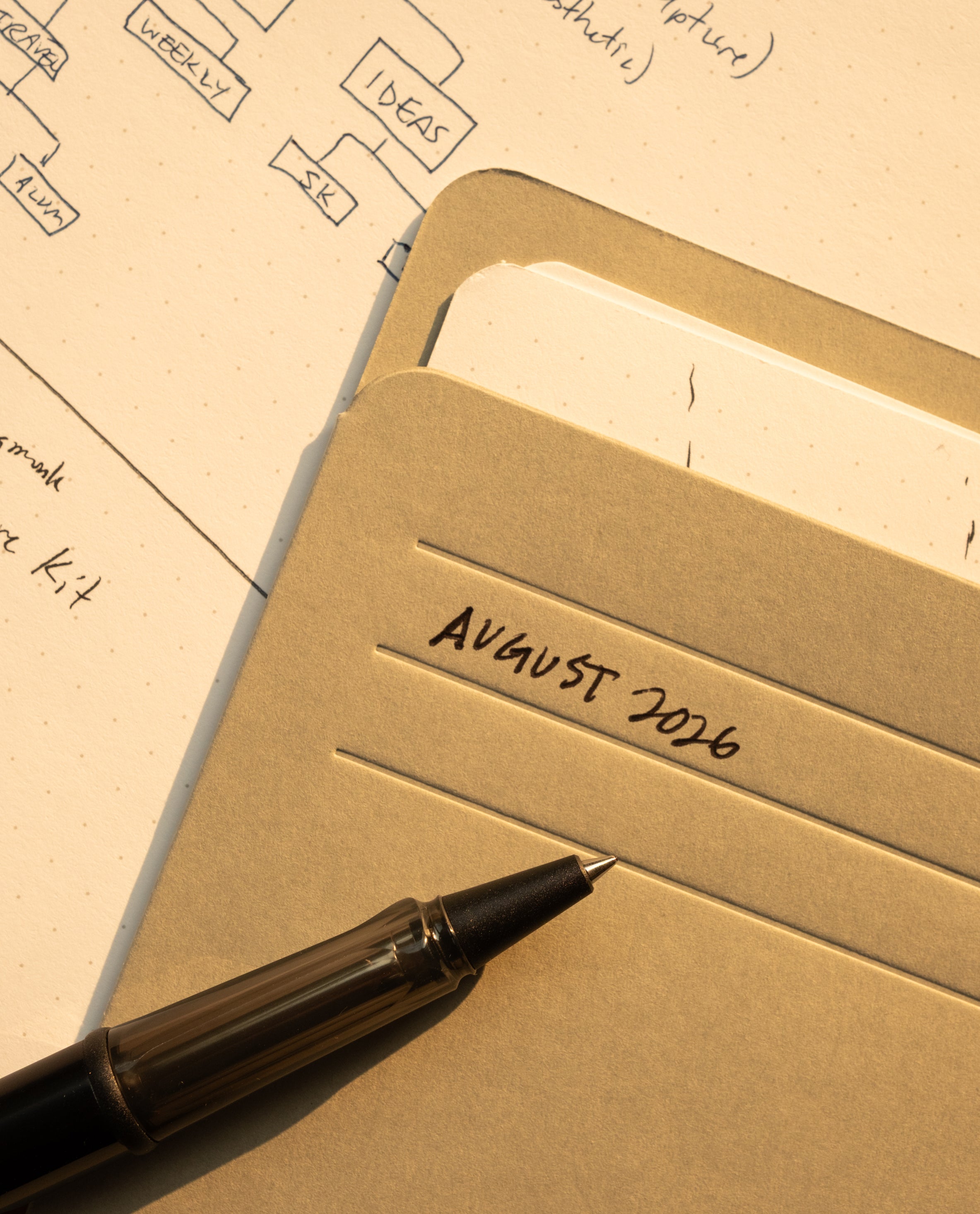 A black pen rests on a folder labeled AUGUST 2026 with papers inside, next to a sheet of paper covered in diagrams and handwritten notes, all illuminated by warm light. Nearby, a sleek card organizer filled with Analog Card Sleeves - Large (Monochromatic) adds an extra touch of order to the workspace.
