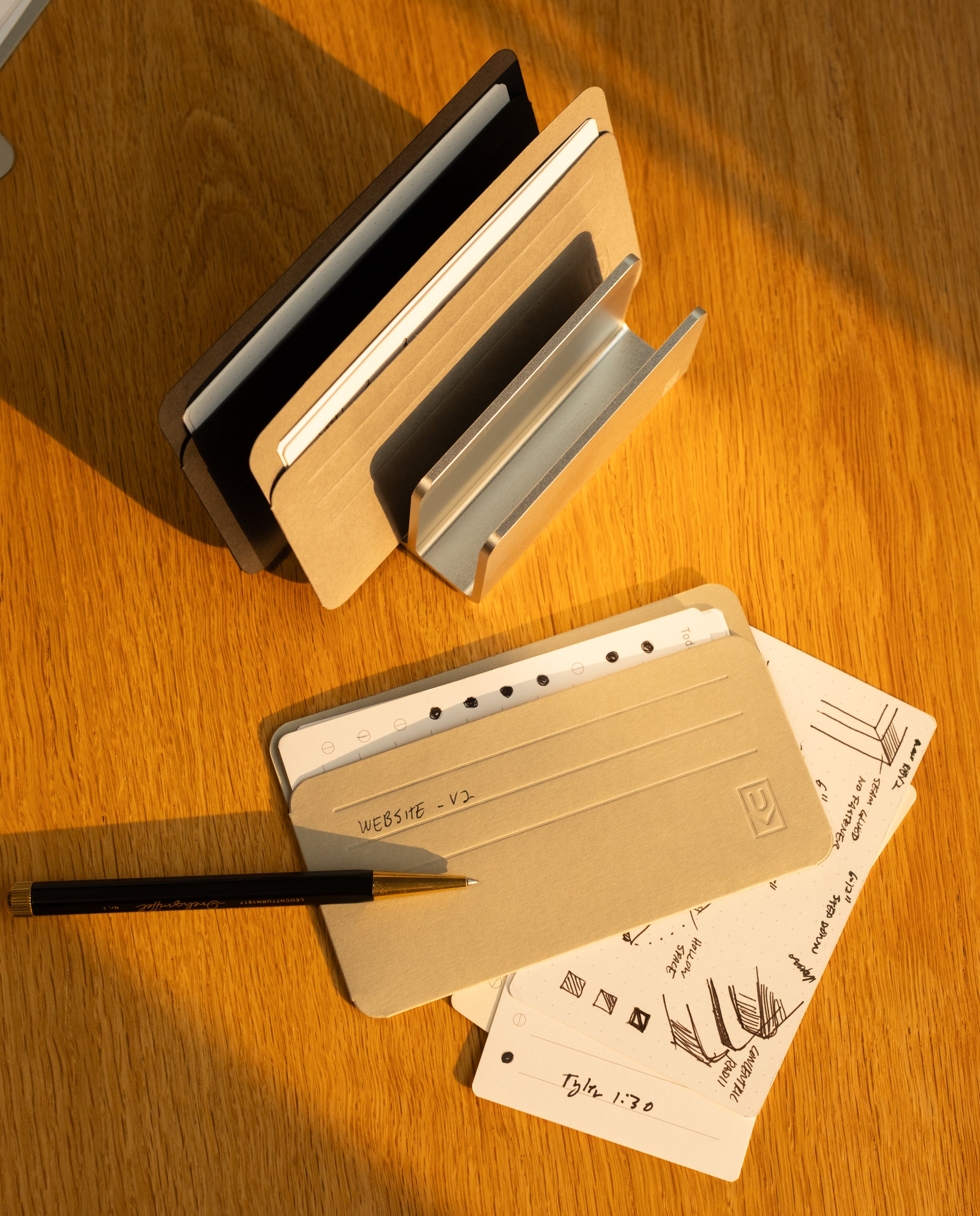 A desk organizer holds notebooks. On the wooden table, a pen rests on a tan notebook. Next to it are open note cards, neatly tucked into Analog Card Sleeves - Small (Monochrome), displaying website sketches and handwritten notes. Sunlight casts warm shadows across the scene.