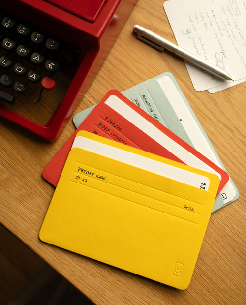 Four colorful index cards labeled with handwritten titles are fanned out on a desk next to a typewriter, a pen, and a handwritten note. The closest card is yellow and titled PROJECT IDEAS 01-12. Each card is neatly stored in its own Analog Card Sleeves – Large (Bright), creating an organized and stylish card organizer for all your project inspirations.