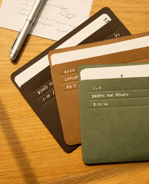 Three labeled file folders—brown, black, and green—are arranged on a wooden desk beside a silver pen and a piece of paper with handwritten notes. The folders, marked with different project names and dates, are neatly stacked next to a compact card organizer filled with Analog Card Sleeves - Large (Earth) for keeping important project cards secure and accessible.