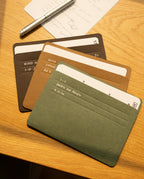 Four colored card holders—Analog Card Sleeves - Large (Earth) in green, brown, and black—are fanned out on a wooden desk near a silver pen and a sheet of notes. These holders are perfect for organizing labeled index cards, ideas, and drafts.