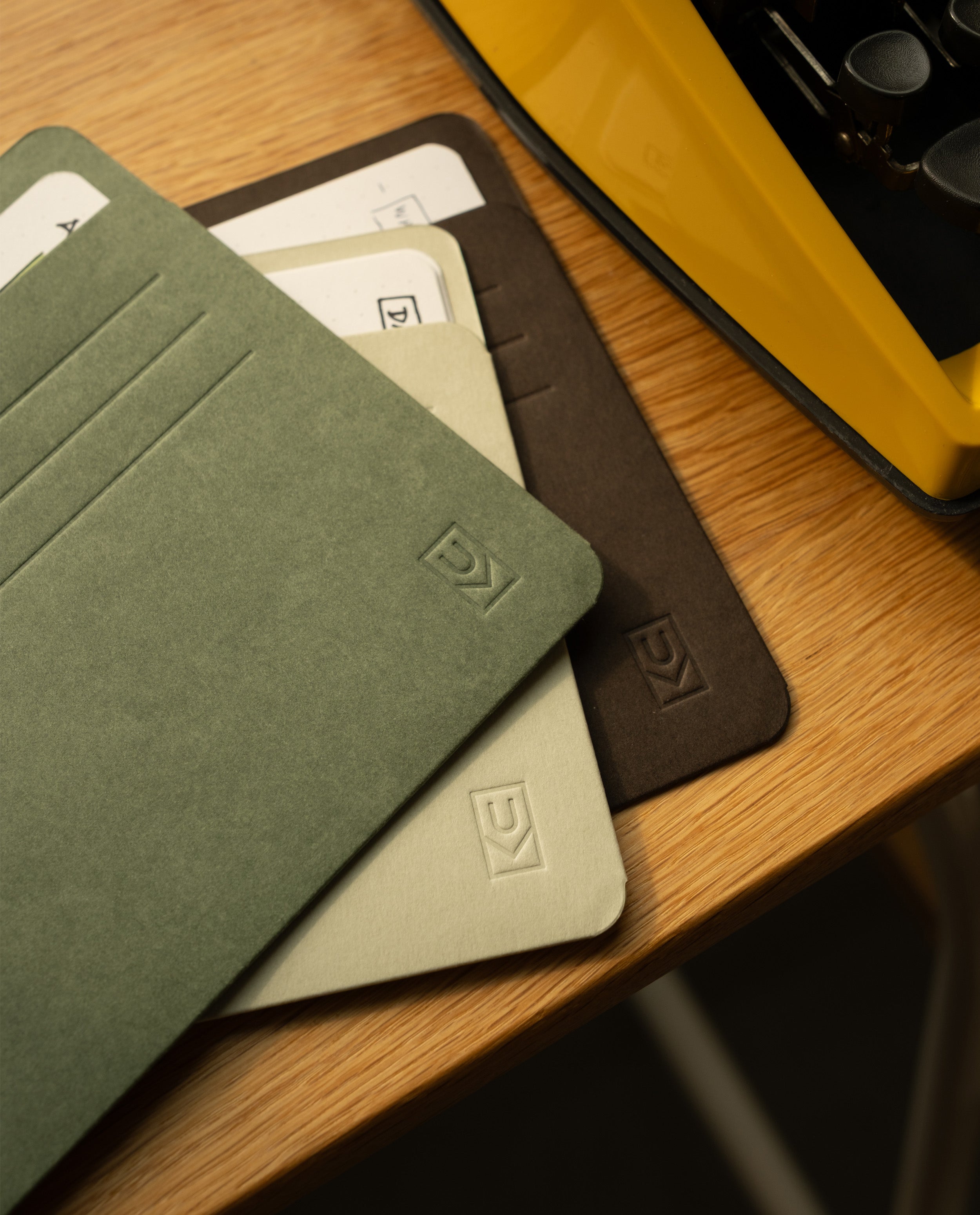 Three Analog Card Sleeves - Large (Earth)—in green, beige, and brown—rest on a wooden desk beside a yellow typewriter. Each sleeve acts as a stylish card organizer with a subtle embossed logo and slots for cards, some of which are partially visible inside. These Analog Card Sleeves - Large (Earth) blend classic design with practical function to keep your essentials neatly arranged.
